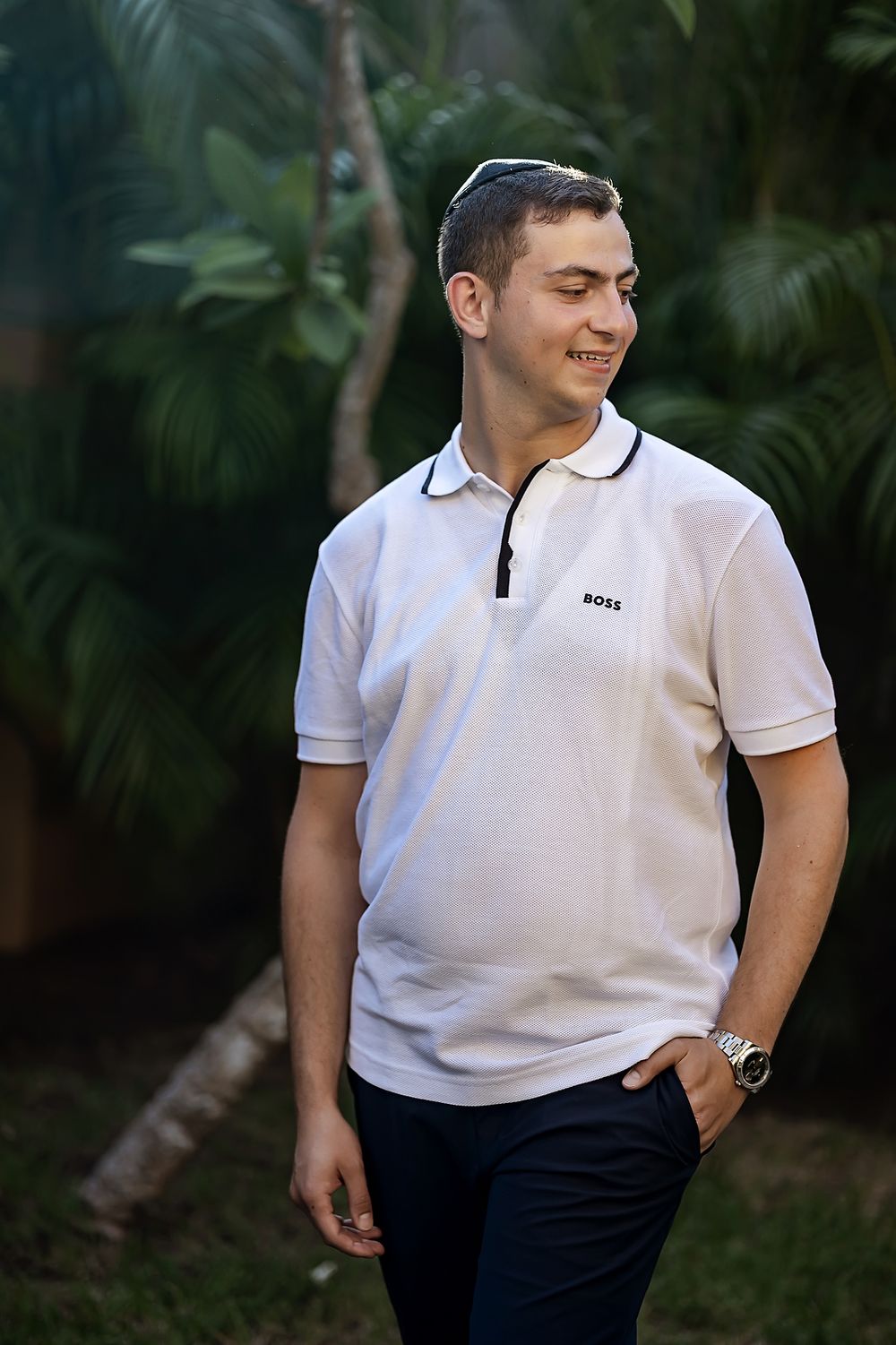 A person in a white polo shirt and yarmulke stands outdoors against a lush tropical garden backdrop.
