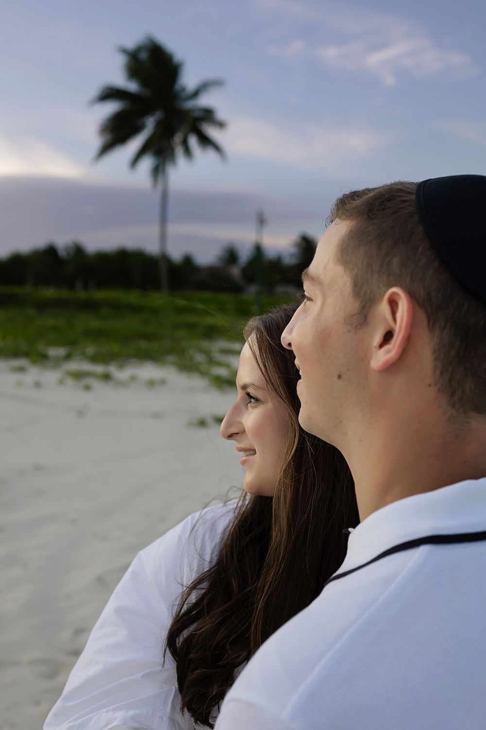 A couple gazes at the sunset on a tropical beach with palm trees in the background.