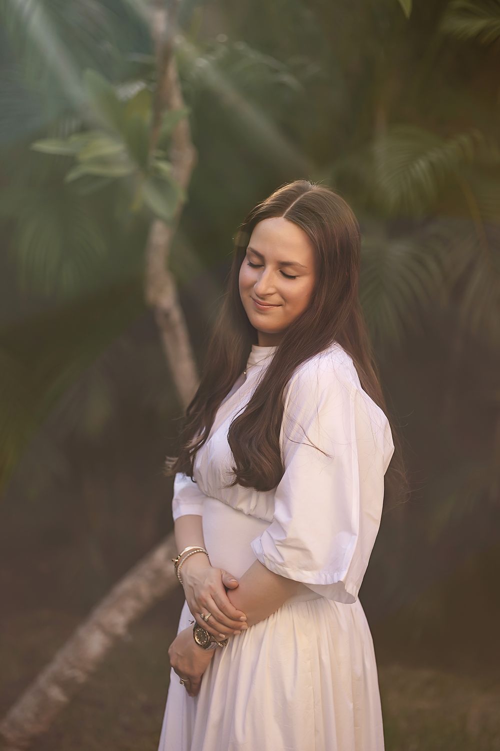 An expectant mother in a flowing white dress poses thoughtfully in a garden setting with soft natural lighting.