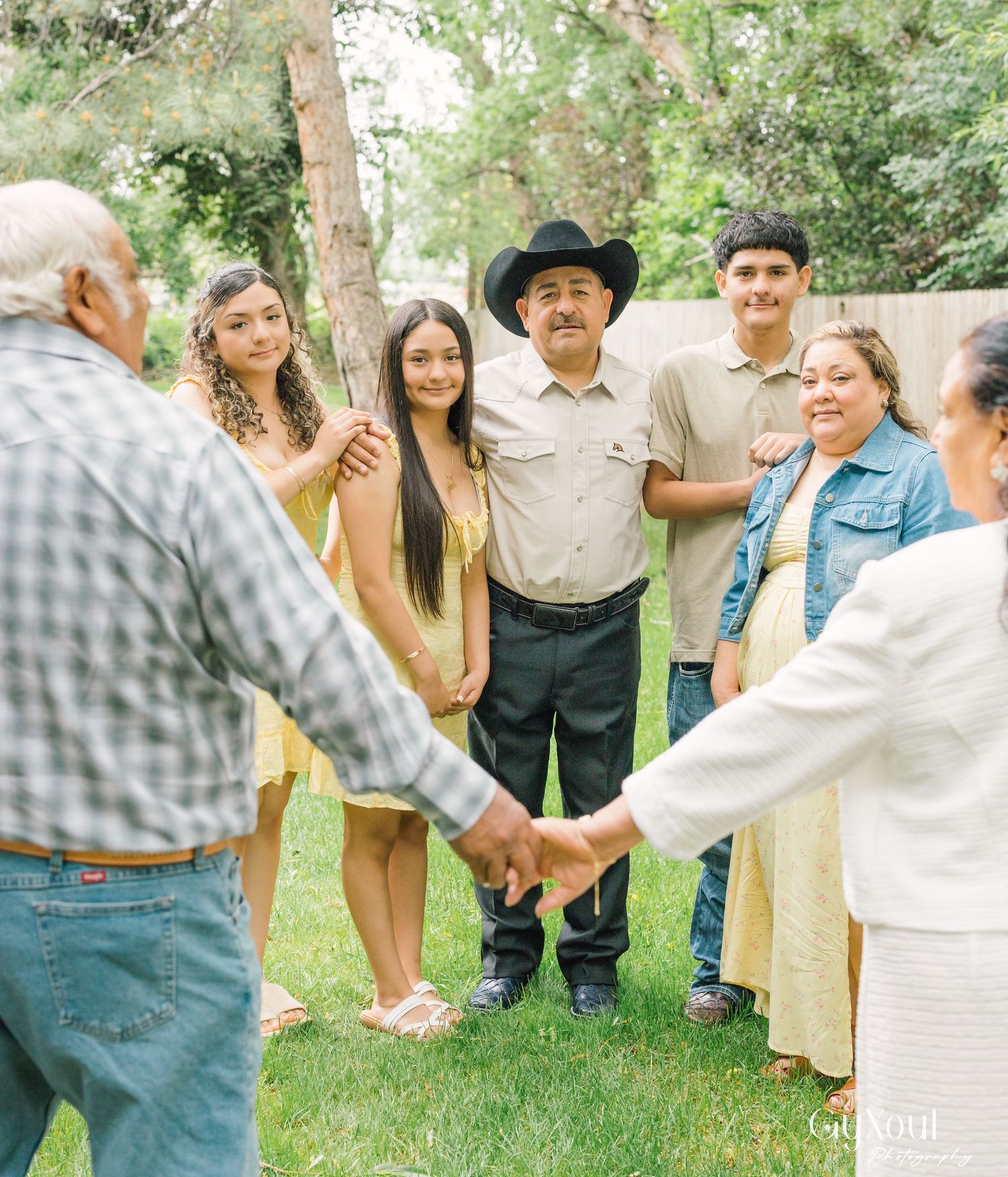 A multi-generational family gathering outdoors in a garden setting while two people hold hands in the foreground.