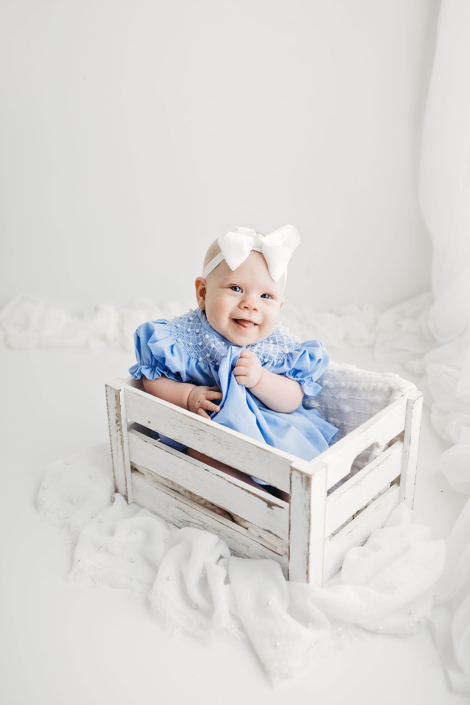 A baby in a light blue dress and white bow sits smiling in a rustic white wooden crate surrounded by white fabric.