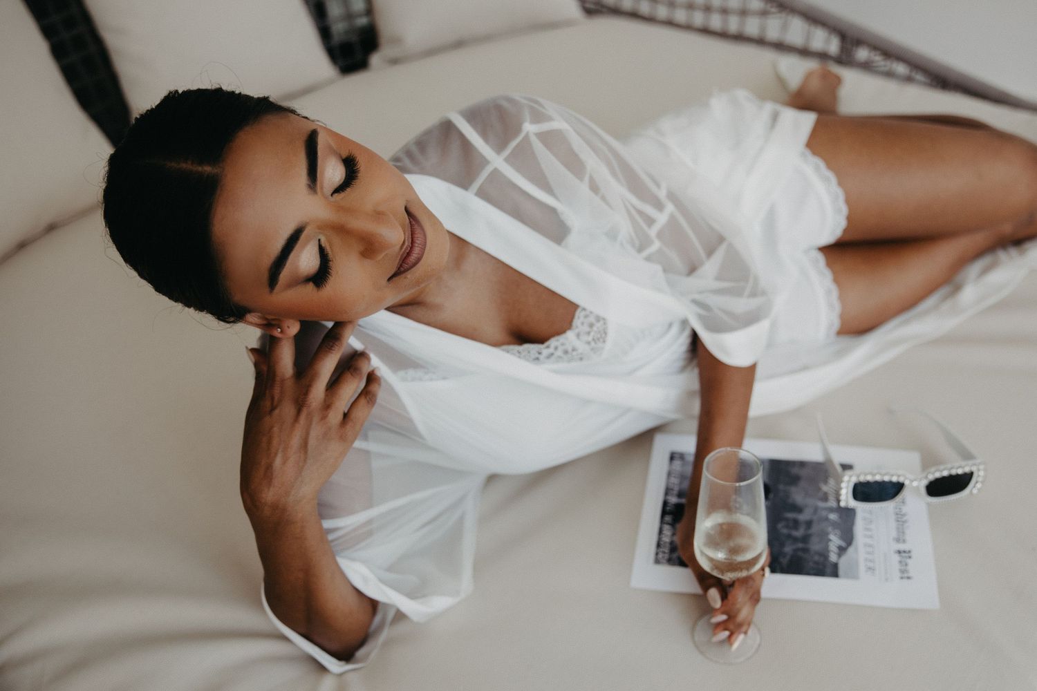 A person in white bridal lingerie poses on a bed during wedding day preparations.