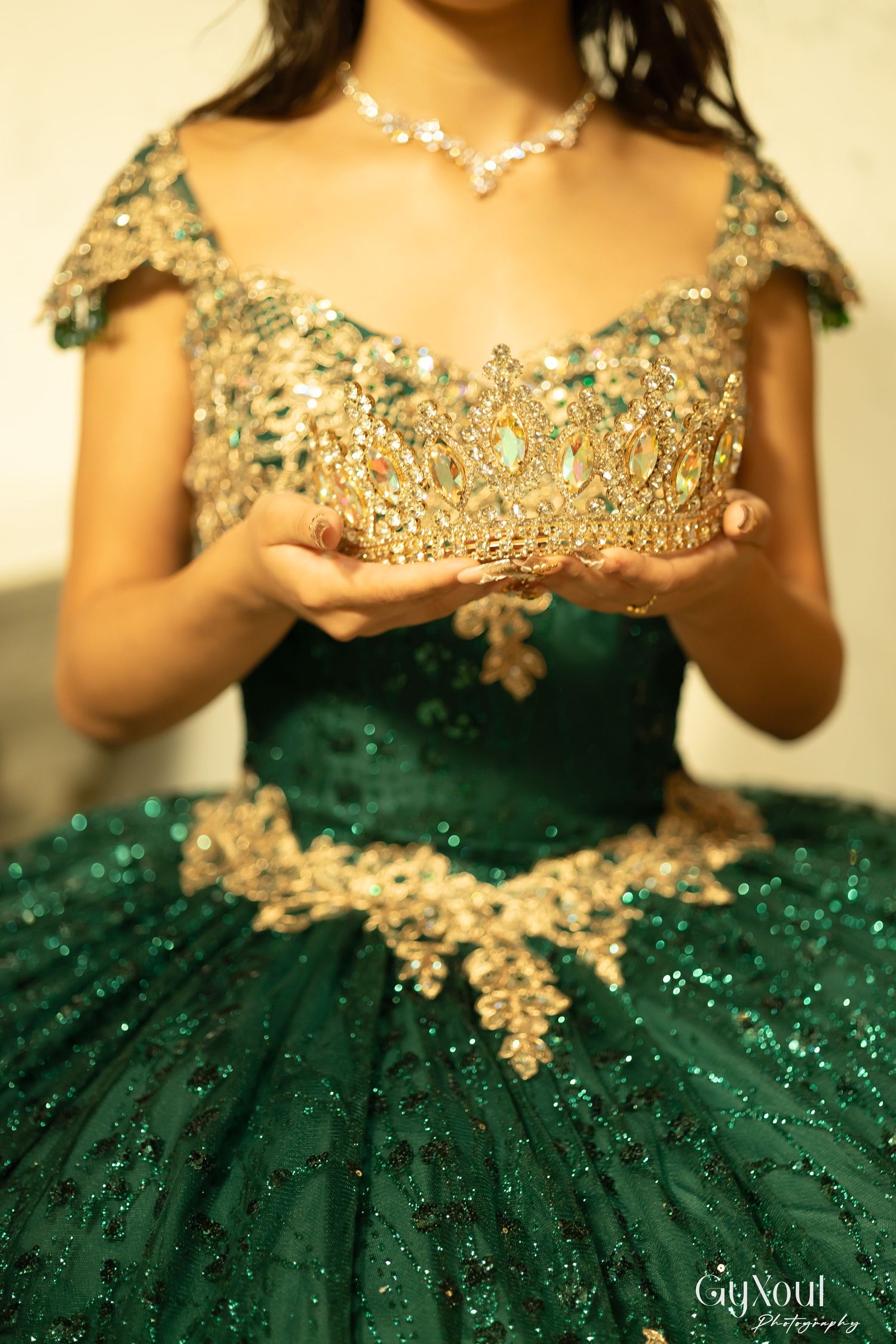 Ornate golden crown held above an emerald green ballgown decorated with intricate gold embellishments and sequins.