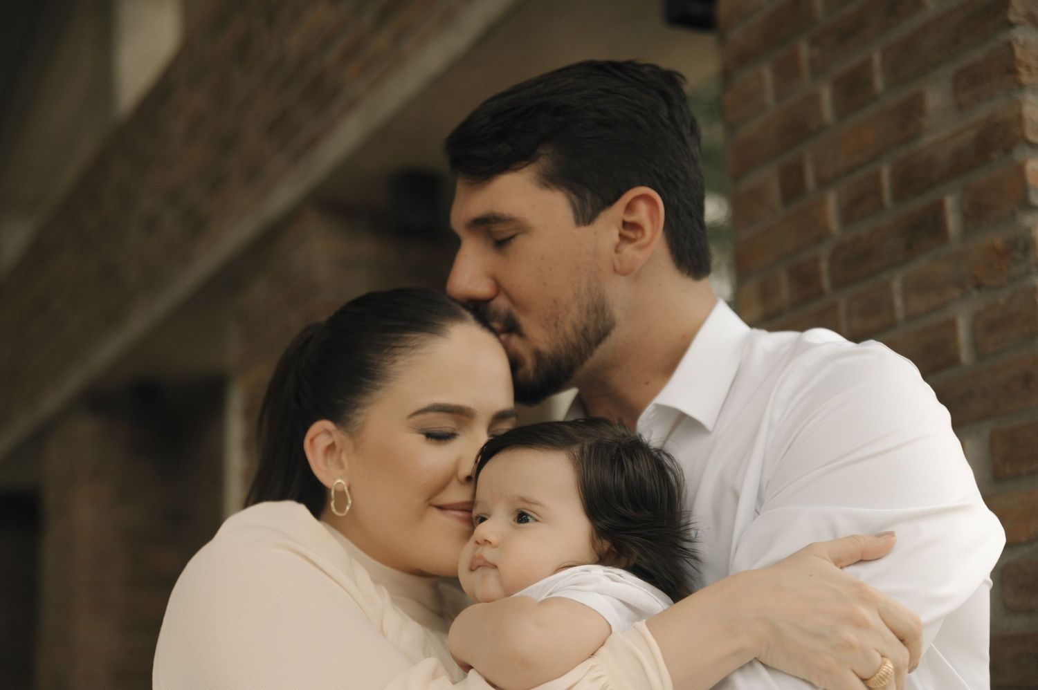 A tender family moment shows parents embracing their young child against a rustic brick wall background.