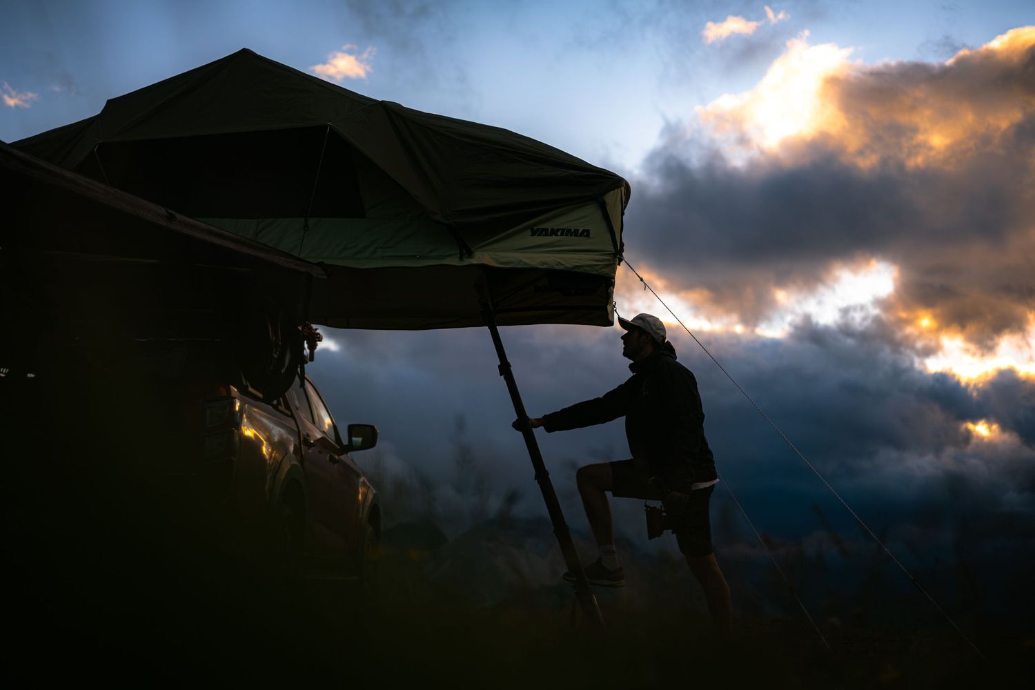 Silhouetted figures and tents against a dramatic sunset sky with dark clouds and golden light.