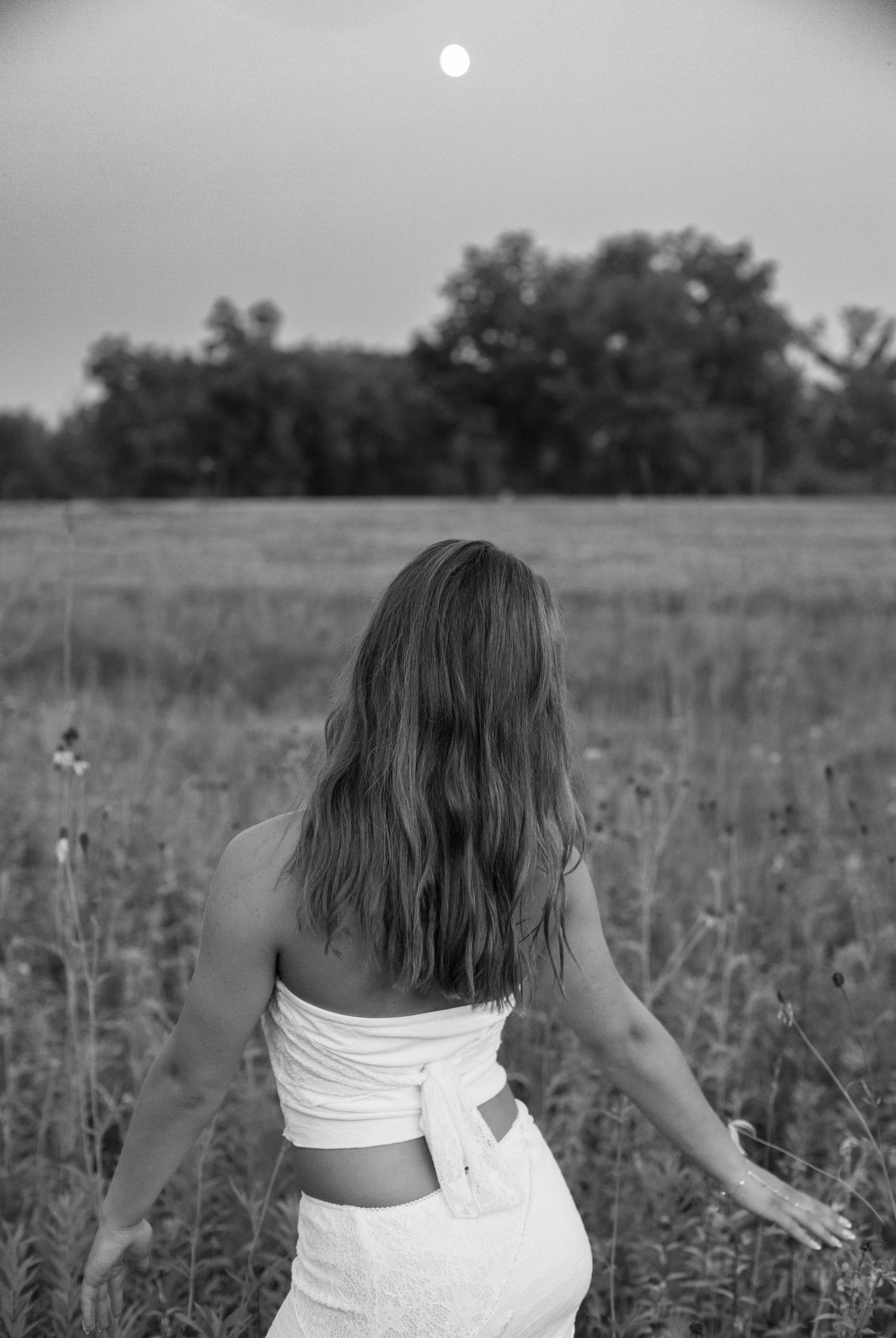 Black and white photo of someone in white summer outfit walking through a field at sunset with trees in background.
