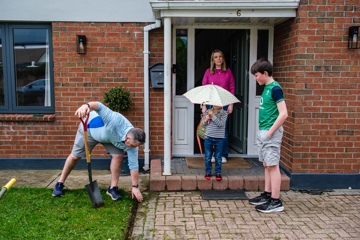People stand in front of a brick house entrance while one person bends down to pick something up from the grass.