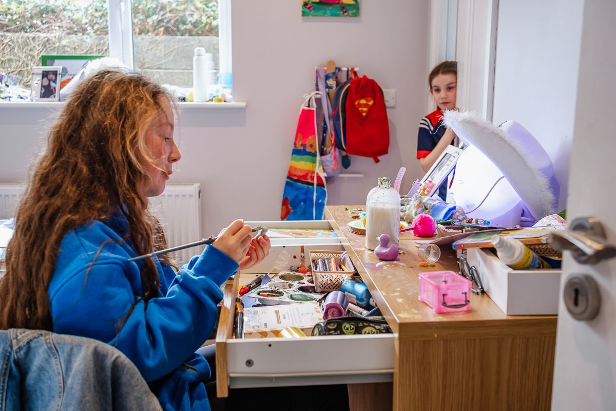 Person wearing blue sweater painting at a craft desk with art supplies and materials spread across wooden workspace.