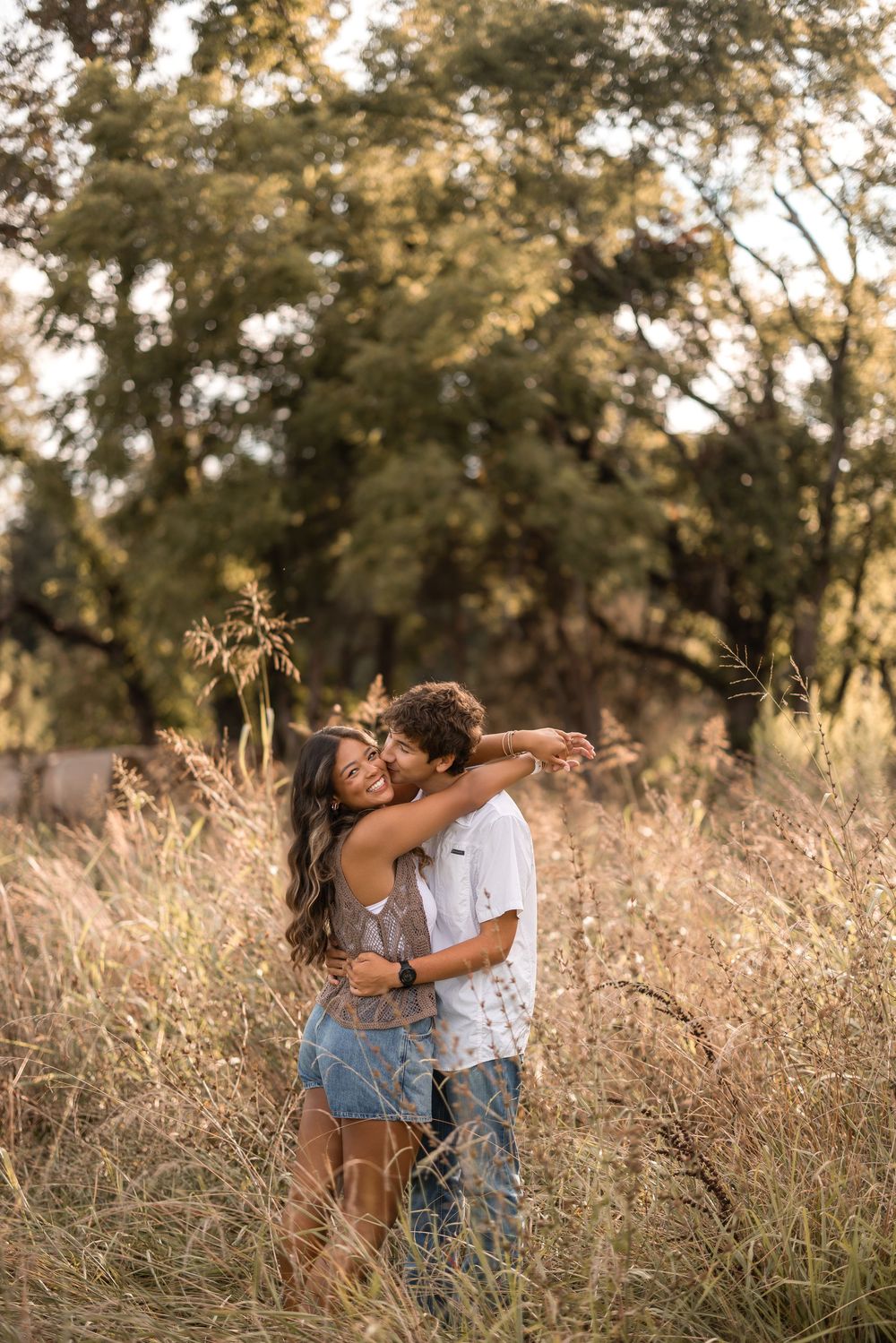 A couple embraces lovingly in a golden meadow during sunset with trees in the background.