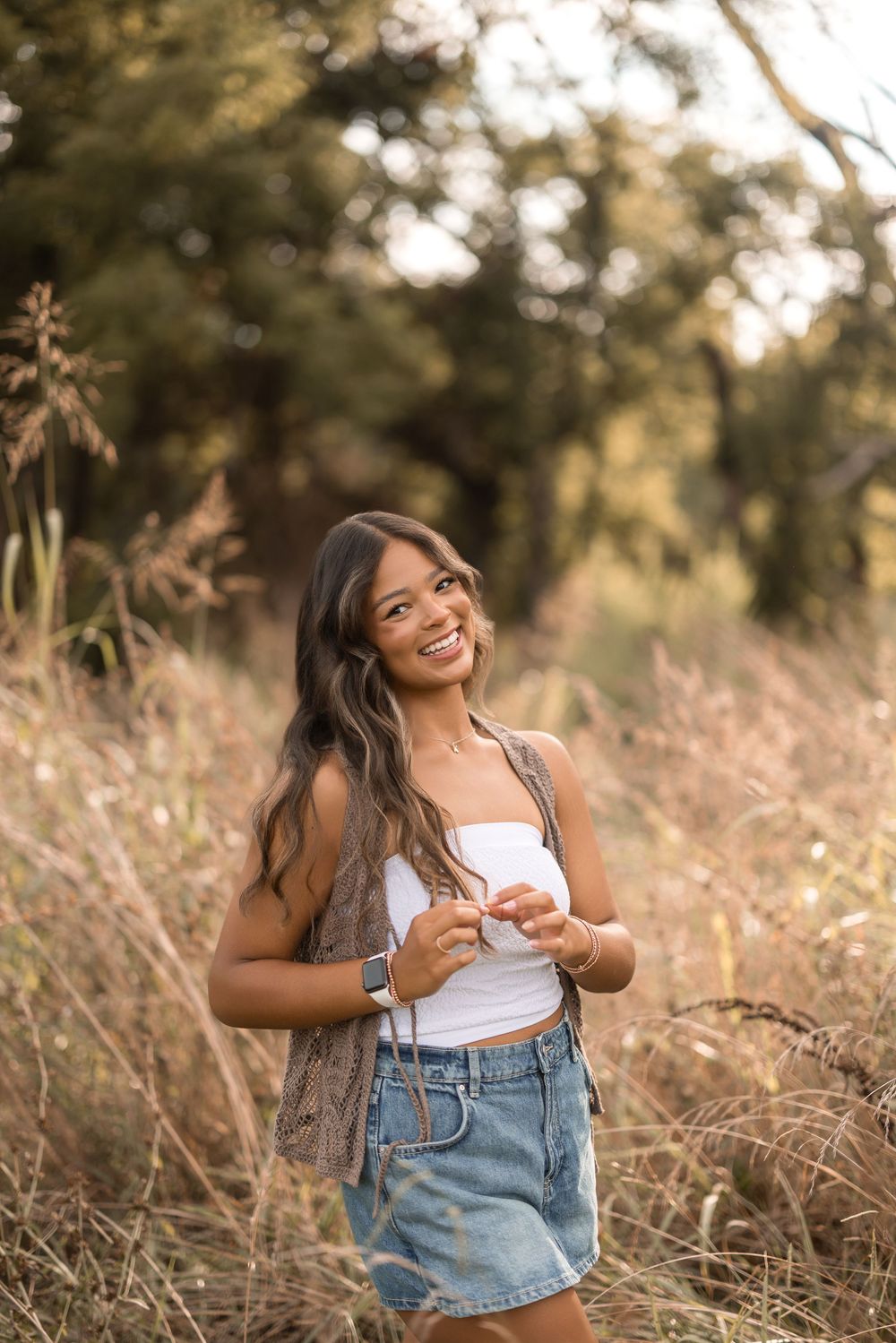 Young woman in denim skirt and white tank top poses outdoors in tall grass during golden hour.