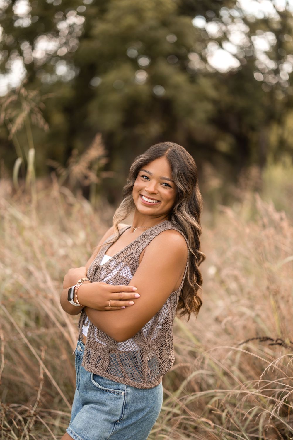Smiling figure posing in field wearing overalls and patterned top against natural bokeh background.