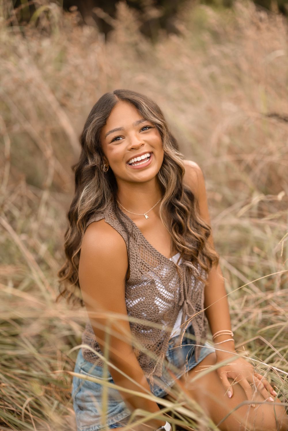 Portrait of someone in a sparkly gold top and denim shorts sitting in tall golden grass at sunset.