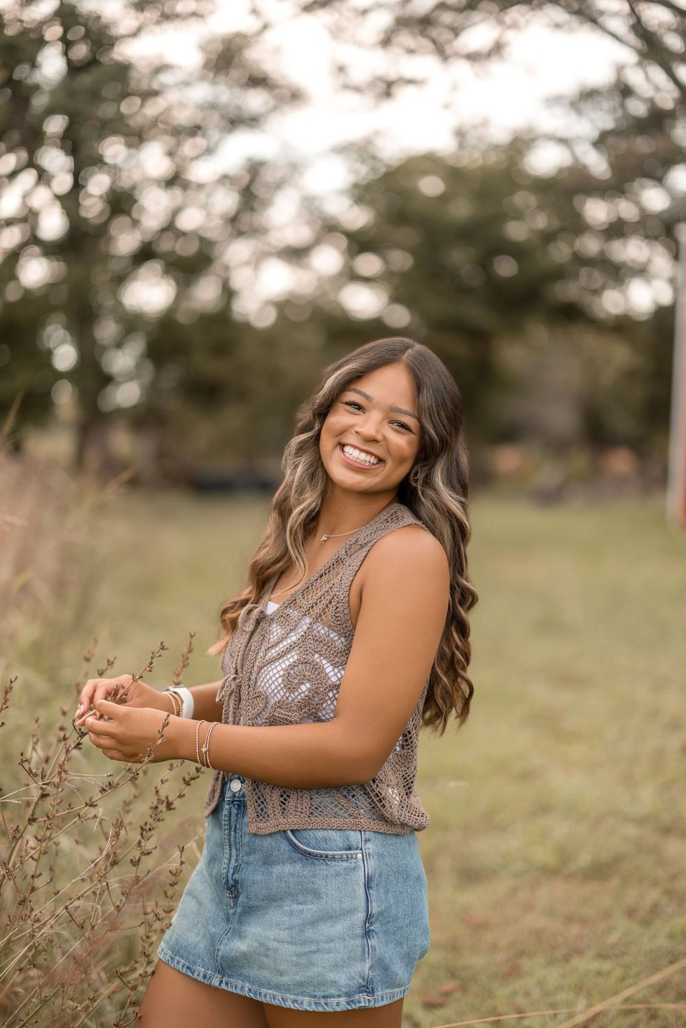 A person wearing a sparkly tank top and denim shorts smiles joyfully in a sunny outdoor setting with bokeh background.