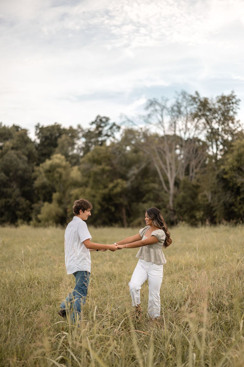 A couple dances and twirls together playfully in a sunlit meadow with tall grass and trees in the background.