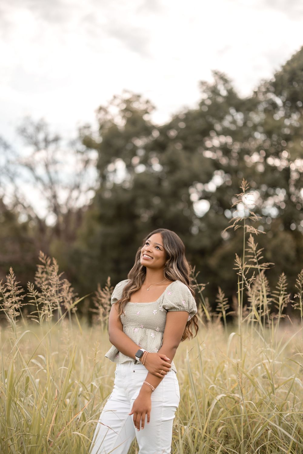 A woman in a white dress stands in a golden wheat field at sunset smiling and looking away from the camera.