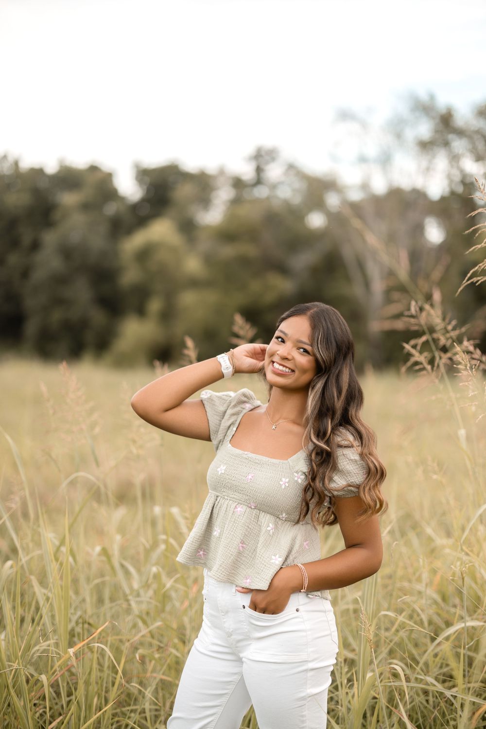 Young woman in white outfit poses cheerfully in tall grass field on sunny afternoon.