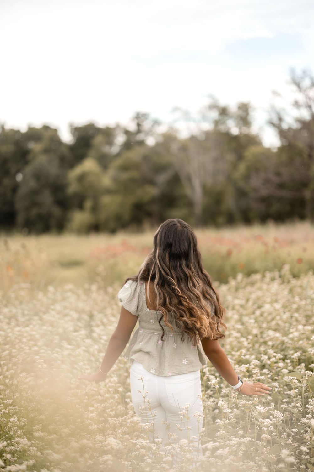 Woman in white dress walking through a field of wildflowers at sunset with tall trees in background.