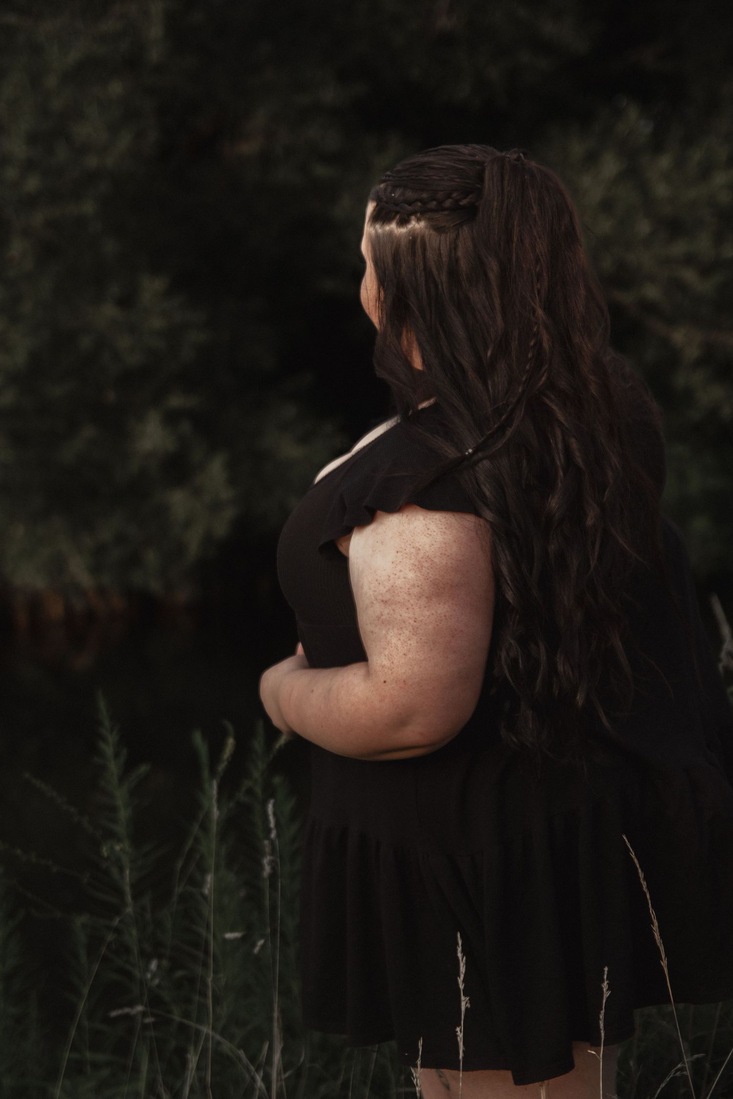 Figure in black dress poses among wild grasses in natural outdoor setting.