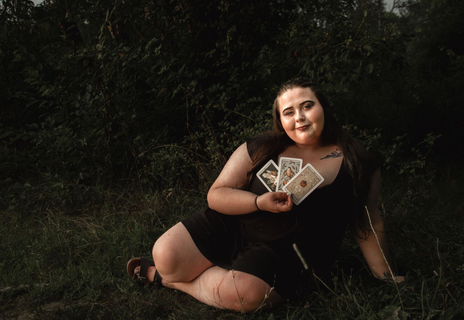 A moody outdoor portrait showing a person in black holding playing cards on grass.