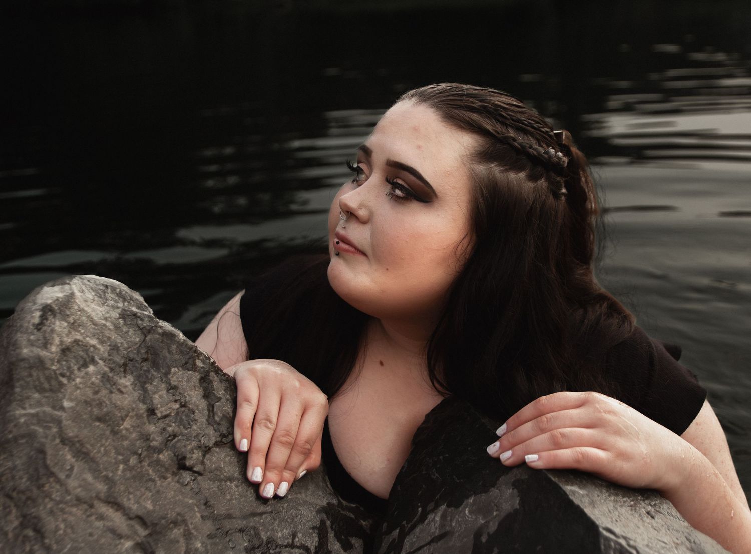 Artistic portrait by a lake featuring dark hair and delicate braids resting against rocks in moody lighting.