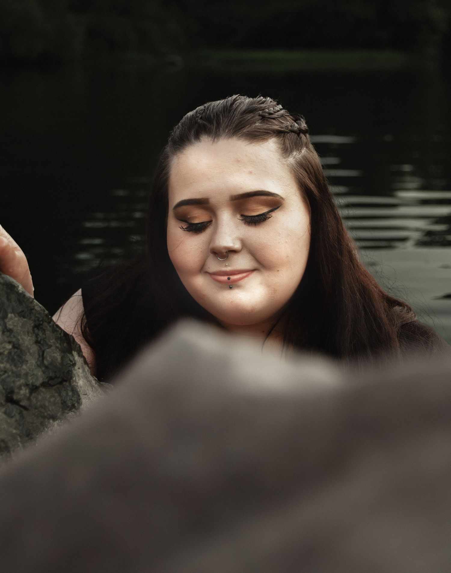 Serene portrait with closed eyes and subtle smile among rocks by water.