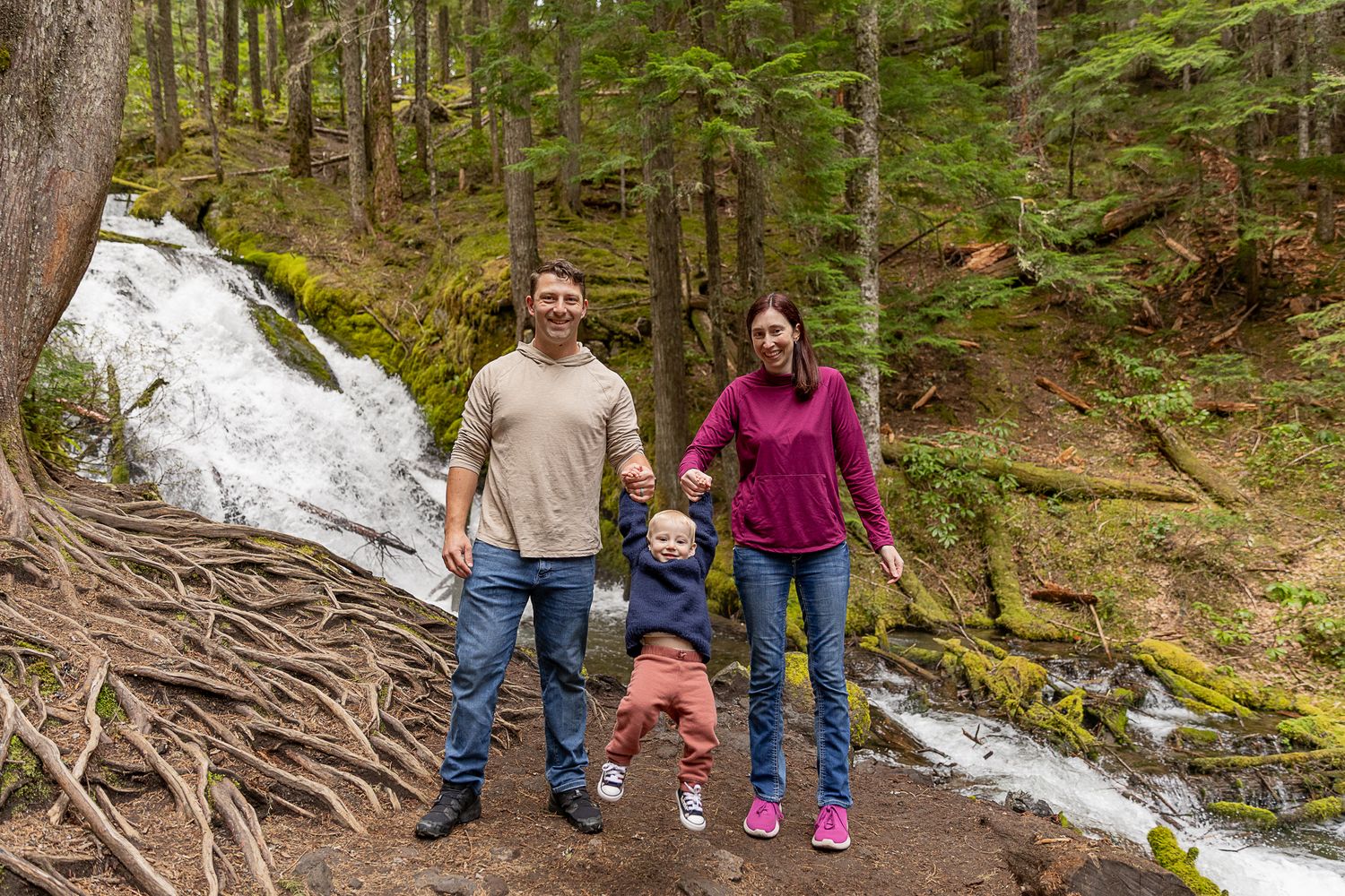 Happy family of three holding hands by a scenic waterfall in a lush forest setting.