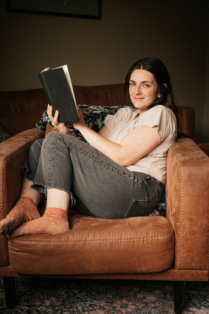 Person relaxing while reading a book on a brown leather couch in a dimly lit room.