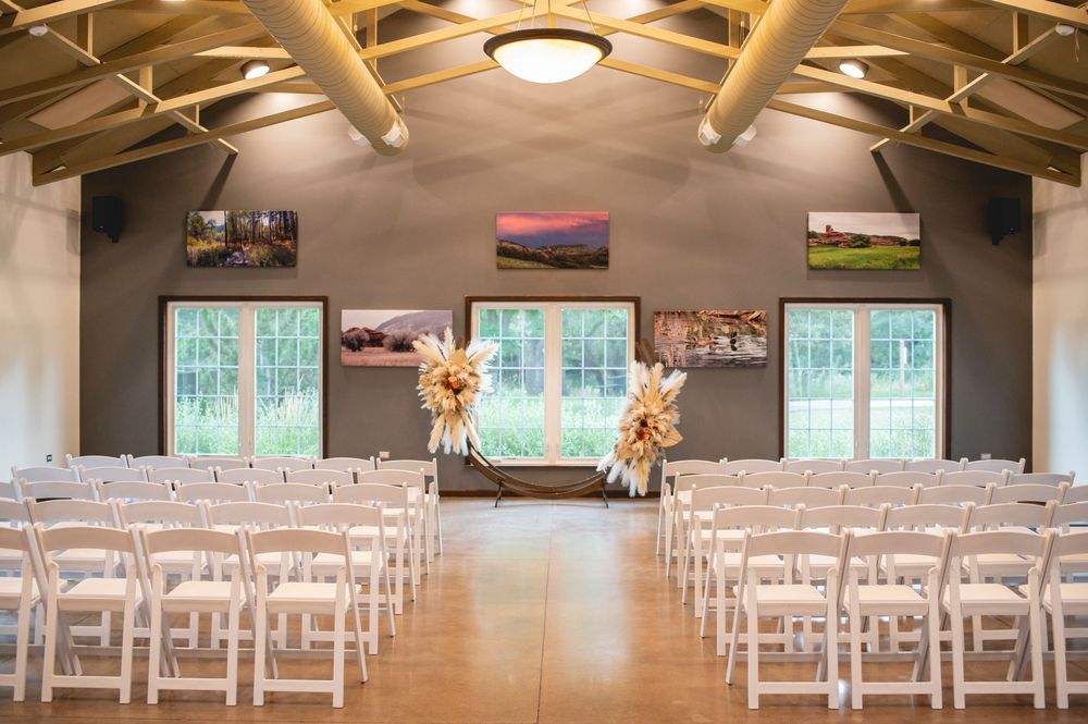 A rustic wedding venue interior with exposed wooden beams, white chairs, and soft lighting for the ceremony setup.