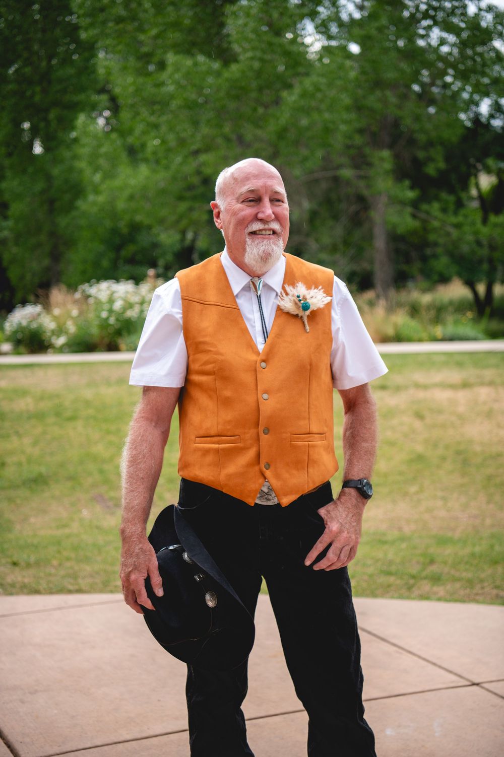 A person wearing a tan leather vest and white shirt with a boutonniere stands outdoors against a green background.