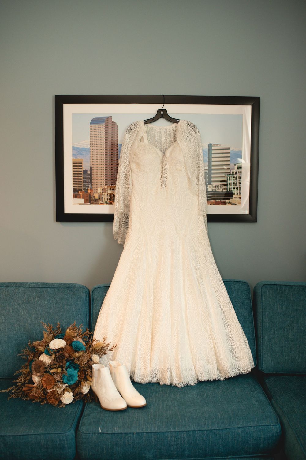 A white lace wedding dress hangs above a teal couch with white boots and a floral bouquet with turquoise accents.