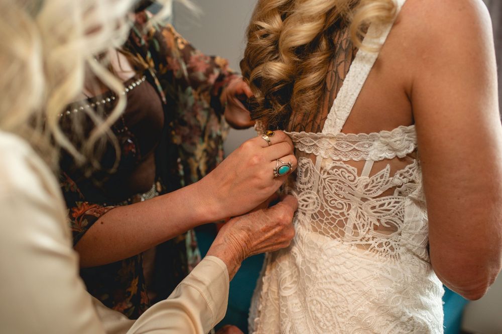 People helping to fasten buttons on an ornate white lace wedding dress during pre-ceremony preparations.