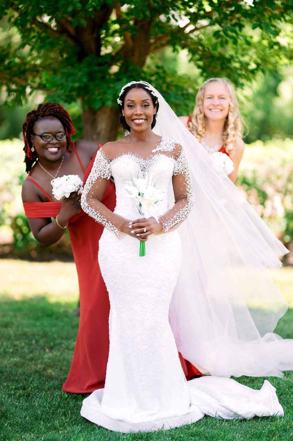 Wedding party poses on lawn with bride in lace gown and bridesmaids in red dresses.