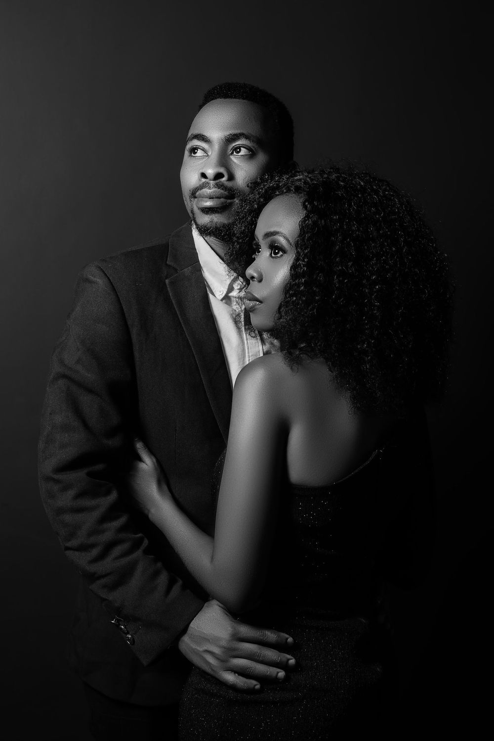 Dramatic black and white portrait of couple embracing in formal attire against dark background.