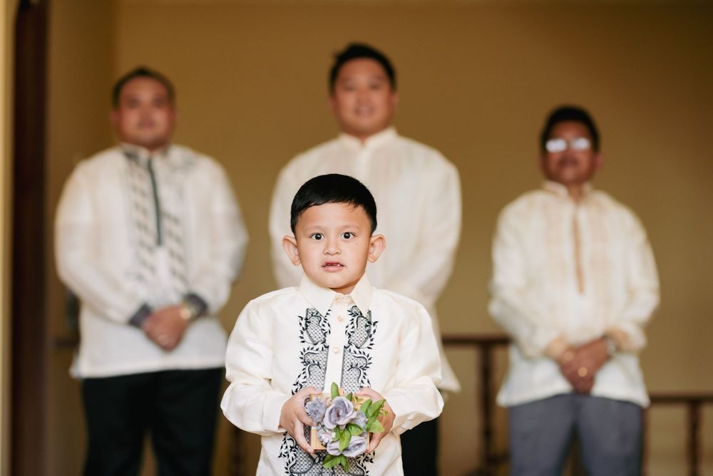 Young ring bearer in traditional white barong tagalog holding flowers with groomsmen in background.