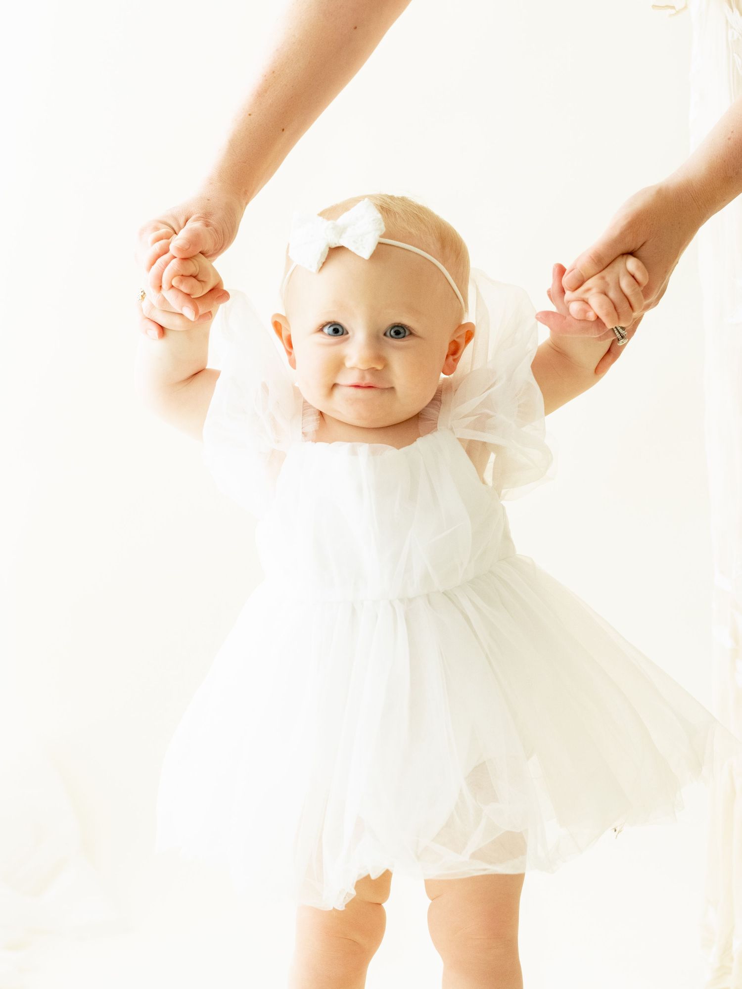 A small baby in a white tulle dress and bow headband holds onto adult hands while standing on a light background.