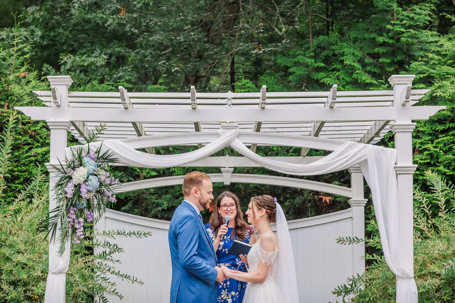 A white wooden wedding arbor decorated with floral arrangements stands in a lush green forest setting.