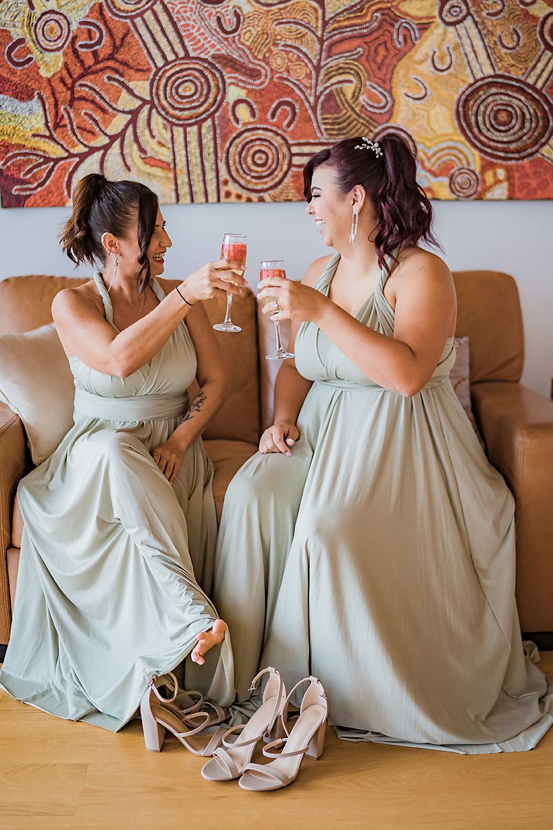 Bridesmaids in sage green dresses toasting with champagne against colorful wall art.