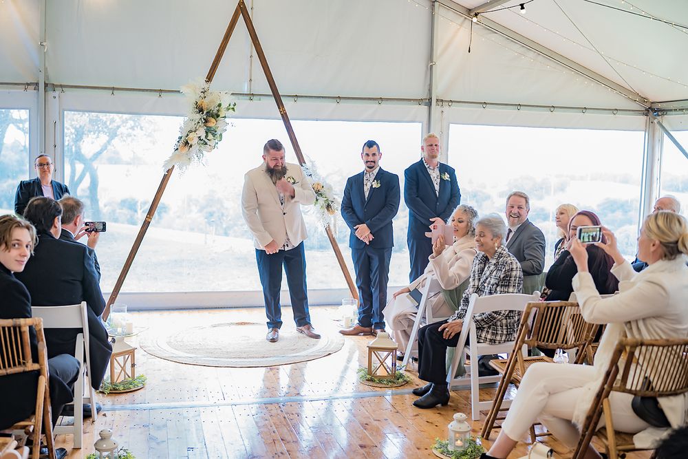 Wedding reception setup in a white tent with wooden chairs and hanging string lights.