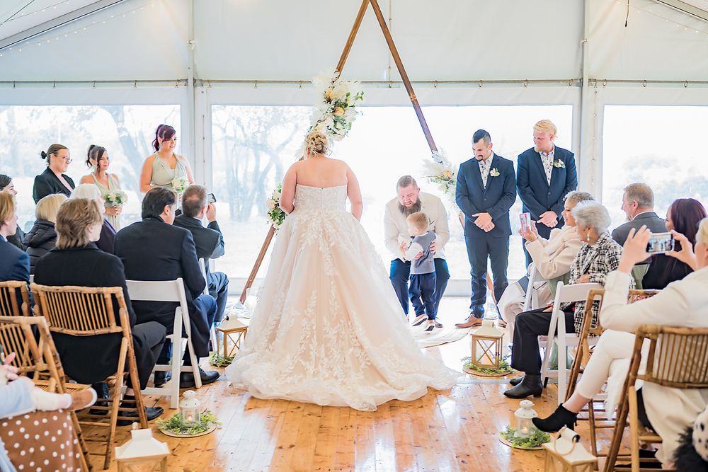 A wedding ceremony takes place under a white tent with guests seated on wooden chairs.