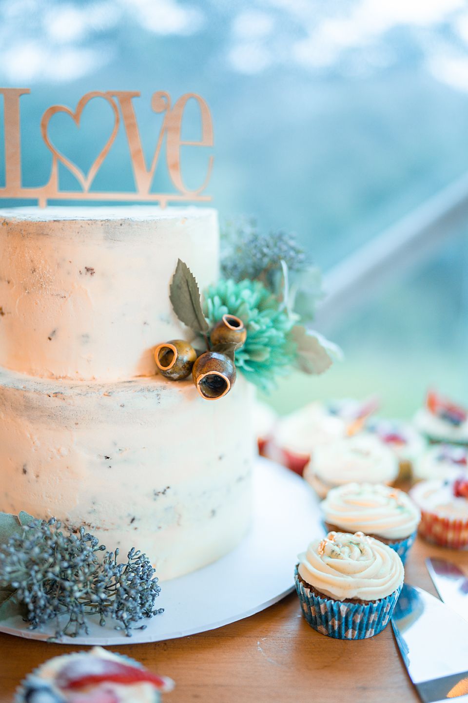 Wedding cake decorated with LOVE topper, dried flowers, and turquoise accents alongside matching frosted cupcakes.
