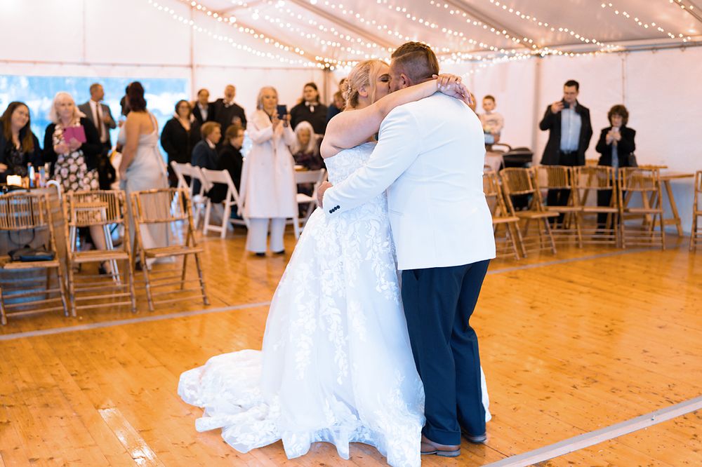 Newlywed couple shares their first dance on a wooden floor under a white tent at their wedding reception.