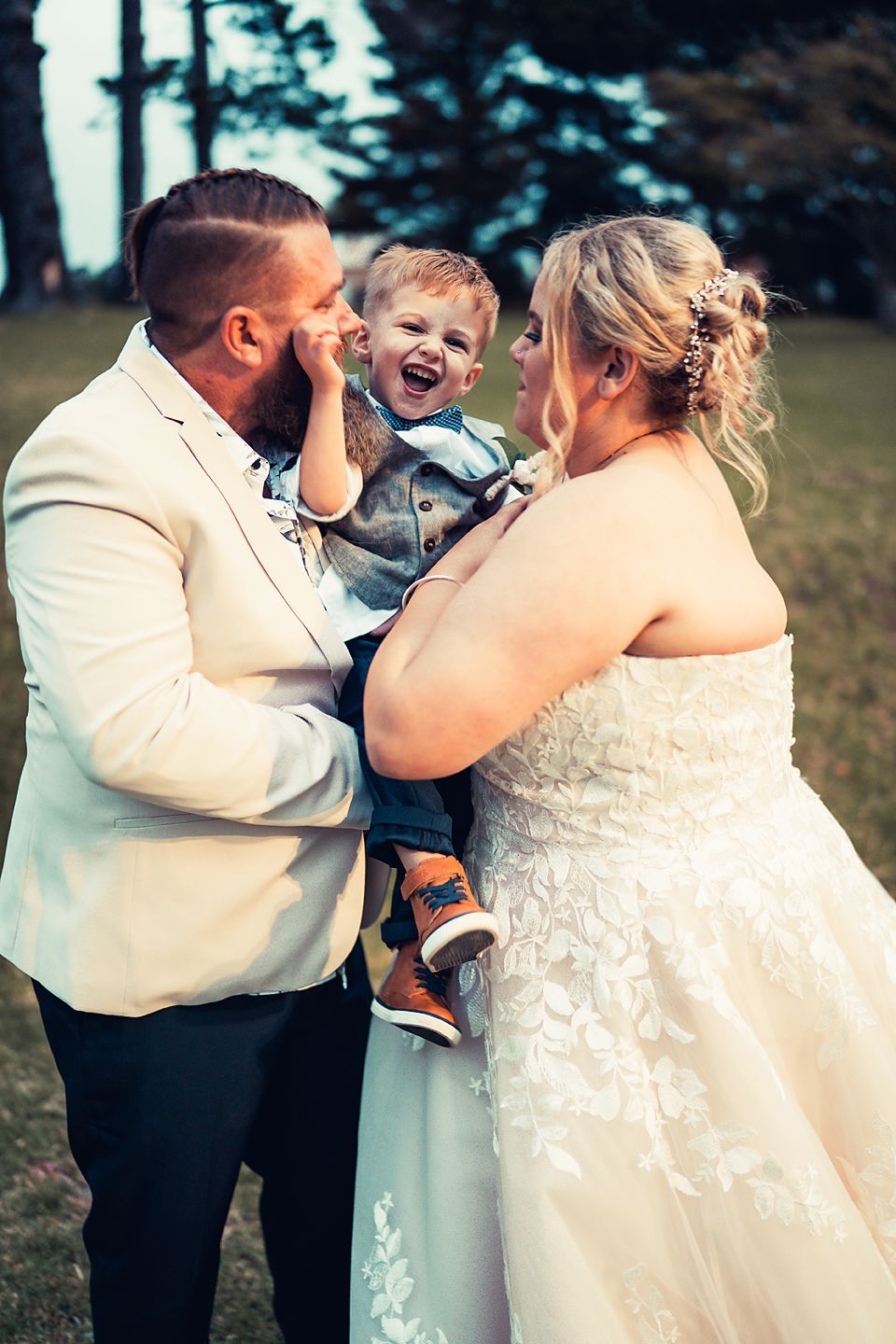 A joyful outdoor wedding moment captured with a laughing child between the bride in white lace and groom in light suit.