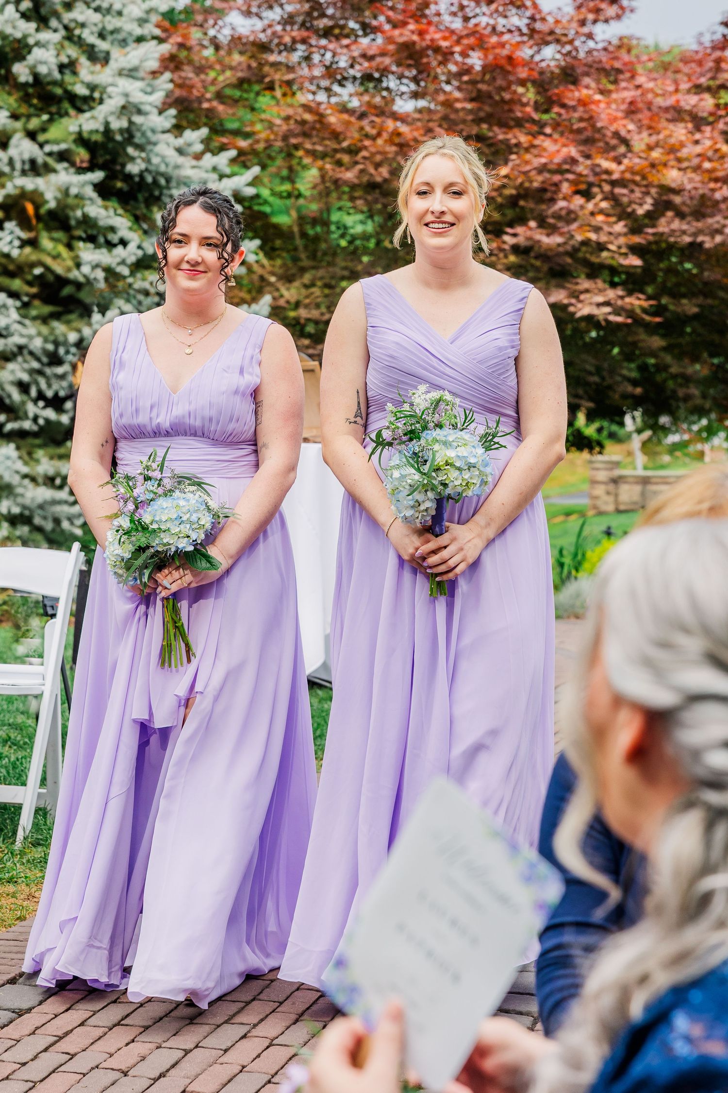 A string of purple-clad bridesmaids holding blue floral bouquets posed together in front of autumn foliage.