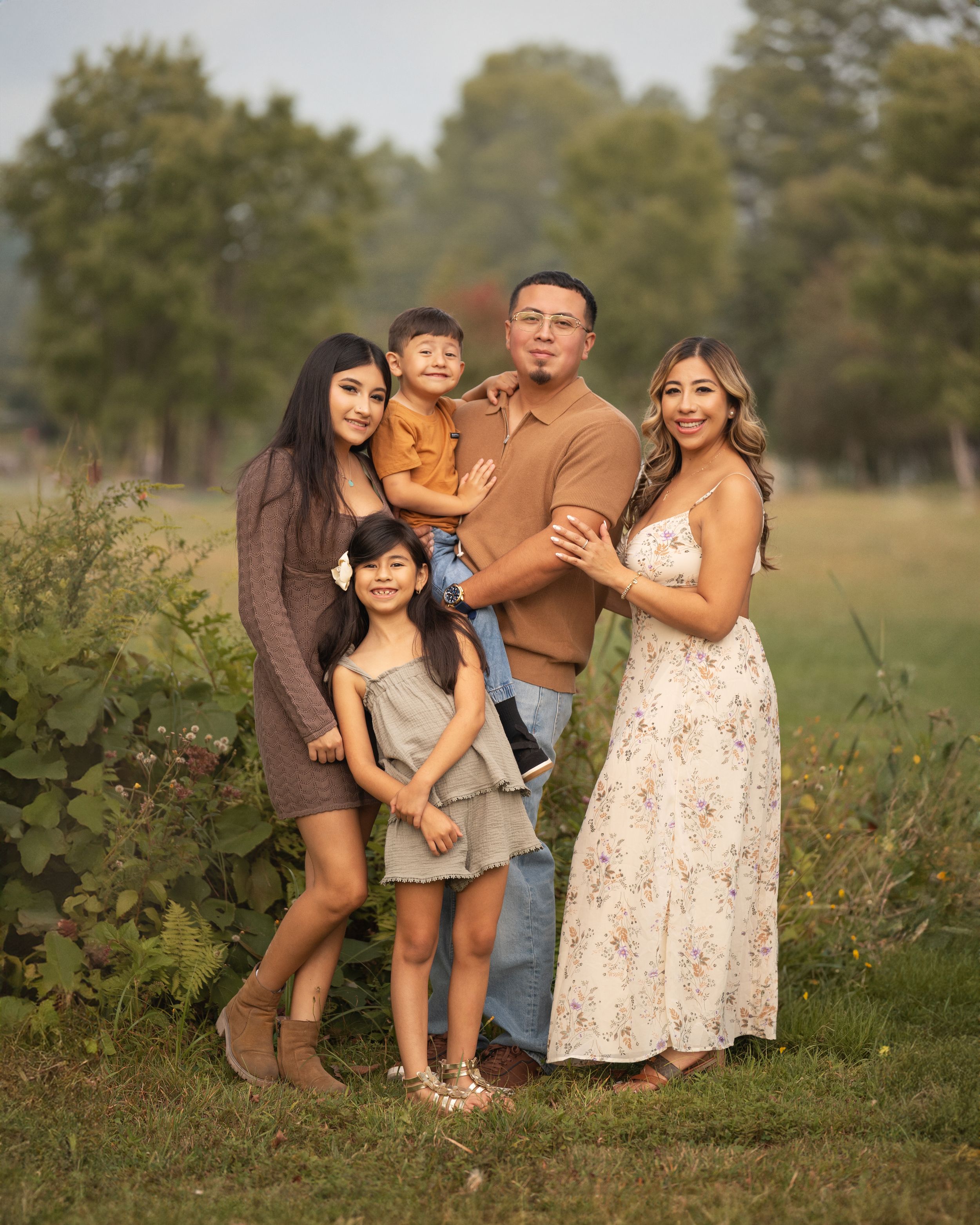 A family poses together outdoors in a field during golden hour, wearing coordinated casual outfits.
