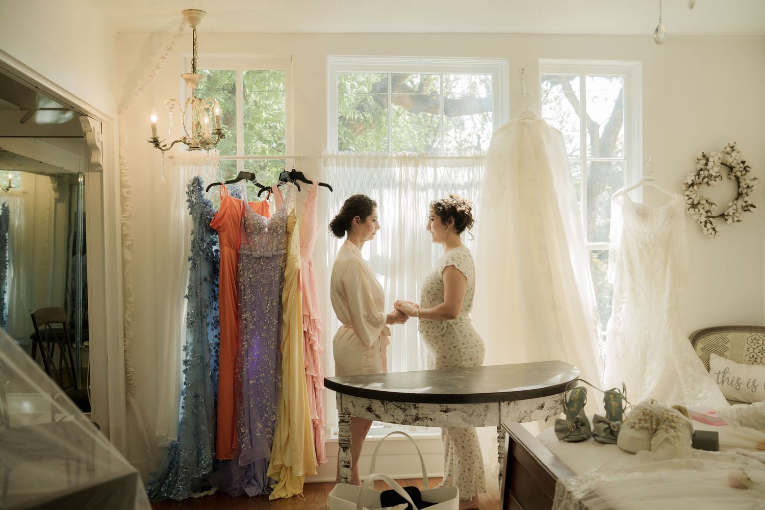 Bridal party preparing in a sun-lit room with colorful dresses hanging.