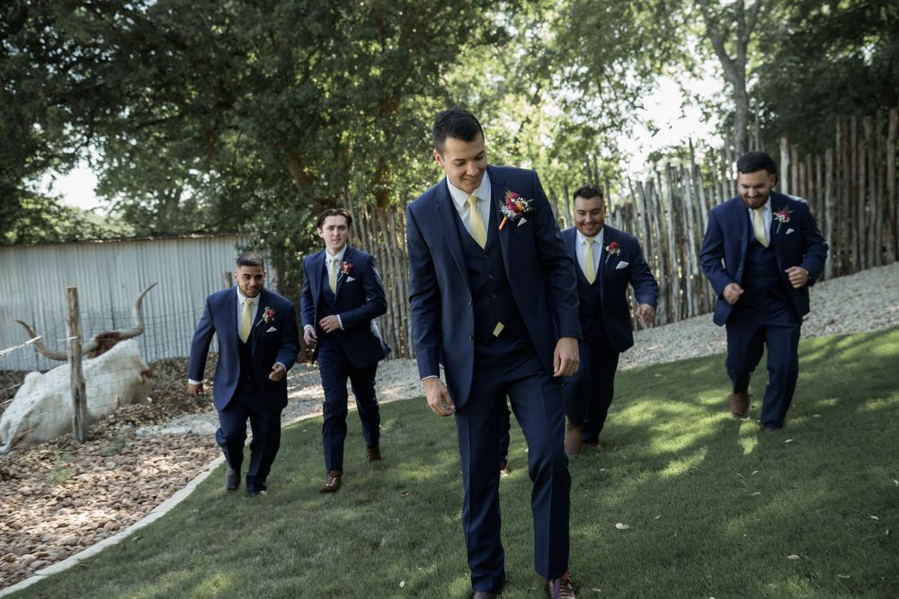 Groomsmen in navy suits walk along a garden path during a wedding celebration.