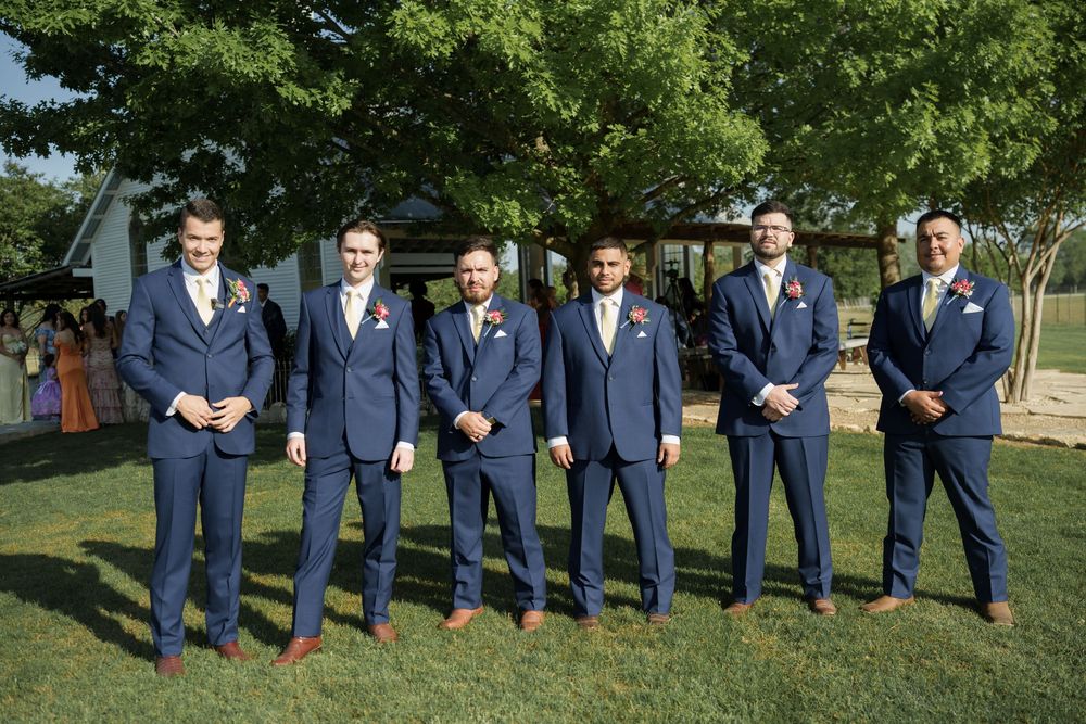 Wedding party in coordinated navy suits poses for formal photo on grass.