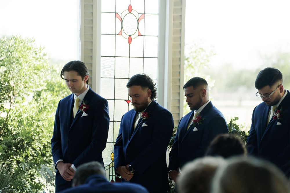 Groomsmen in dark suits bow their heads during wedding ceremony prayer.