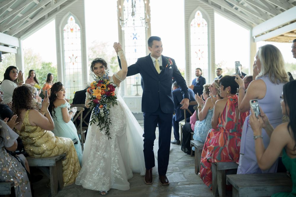 Newlyweds walk down aisle while guests celebrate in sunlit chapel.