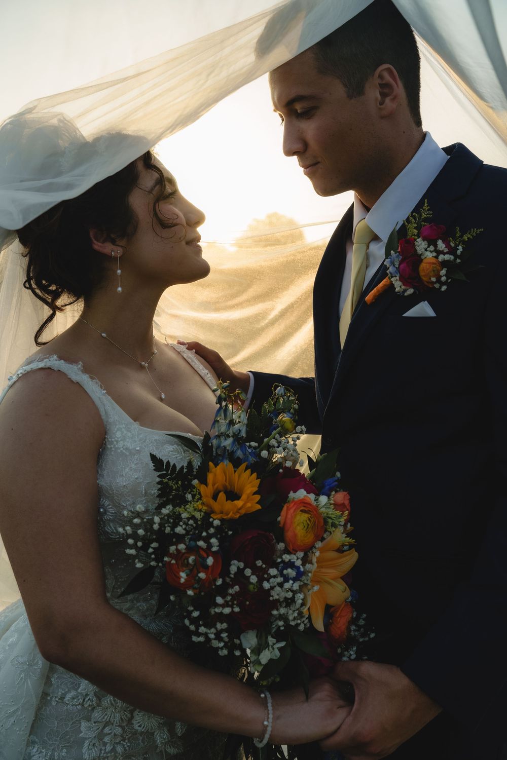 Intimate wedding couple moment with sunflower bouquet and flowing veil at sunset.